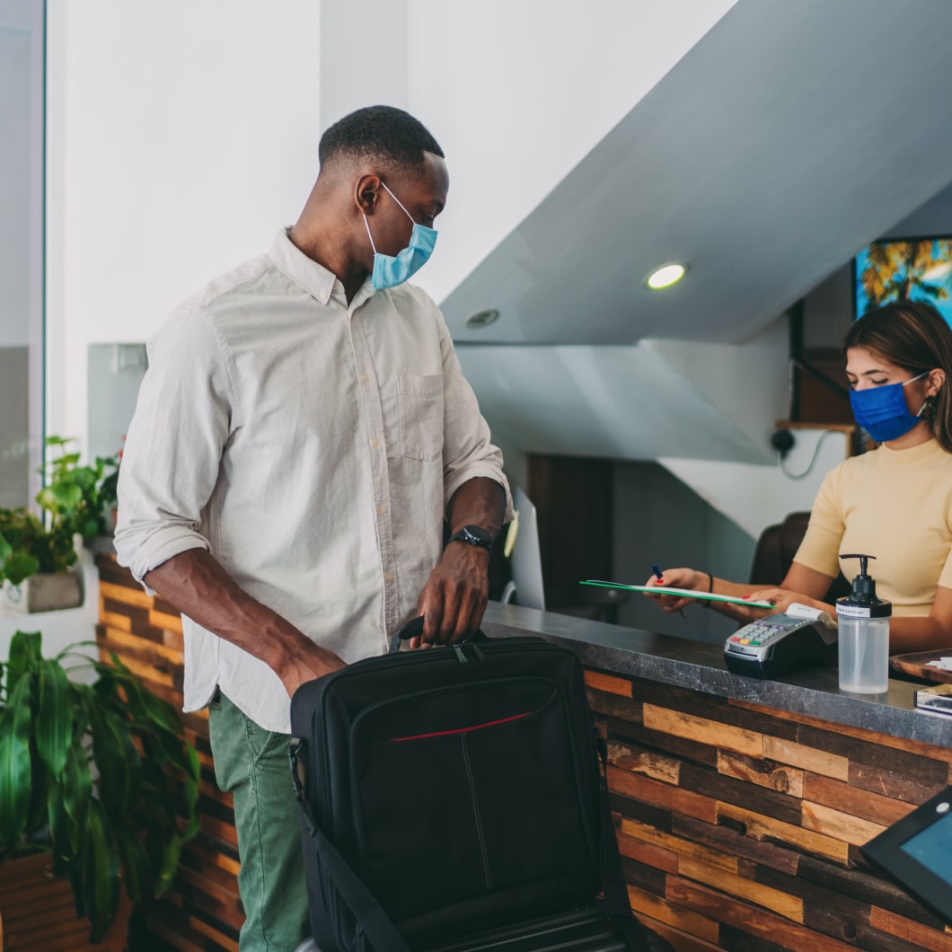 A host checking in a new guest at her hotel, both are wearing masks