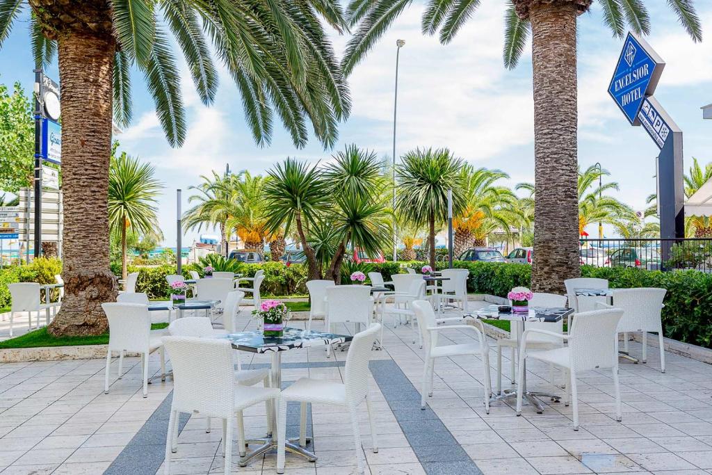 d'une terrasse avec des tables et des chaises blanches et des palmiers. dans l'établissement Hotel Excelsior, à Alba Adriatica