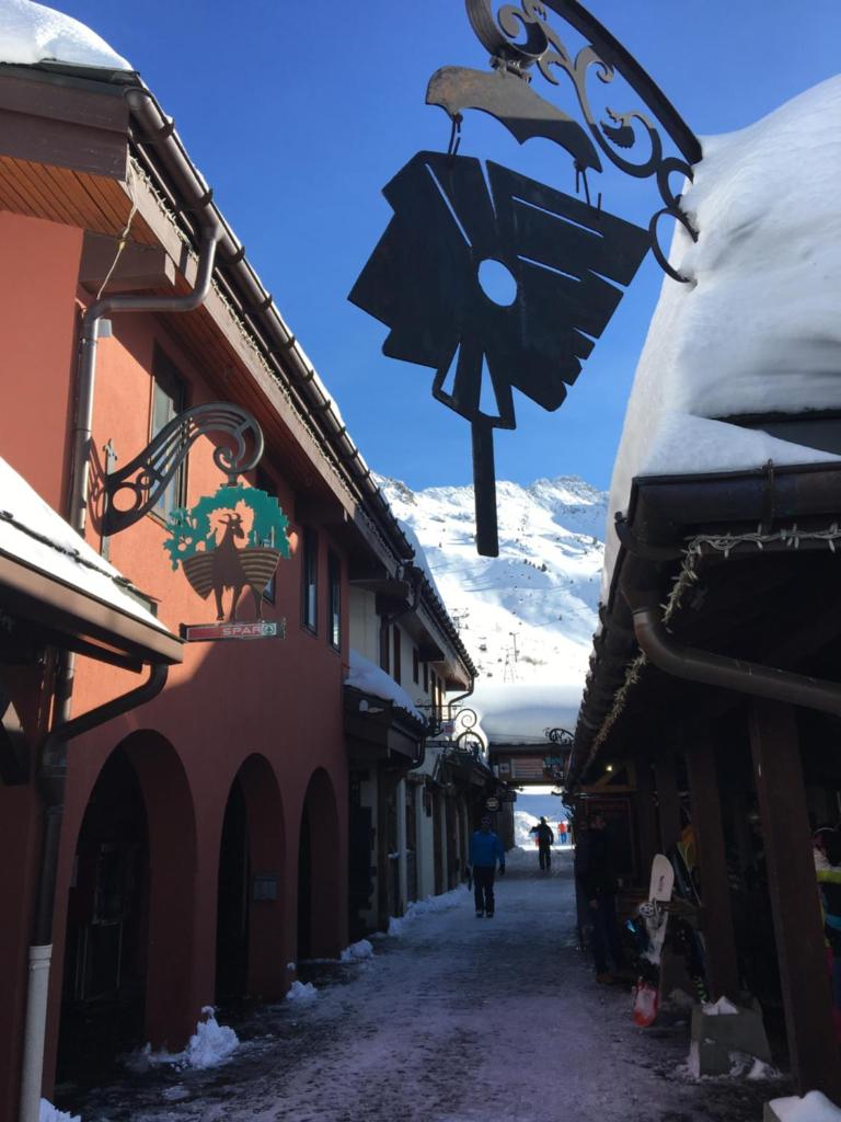une rue avec une horloge sur le côté d'un bâtiment dans l'établissement DUPLEX 6P A BELLE-PLAGNE (2050 m), SKIS AUX PIEDS, EN PLEIN CŒURS DE LA STATION RESIDENCE LE BERYL., à Belle Plagne