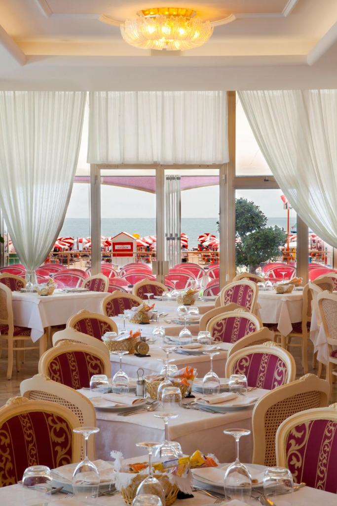 une salle à manger avec des tables et des chaises blanches et des fenêtres dans l'établissement Hotel Excelsior, à Caorle