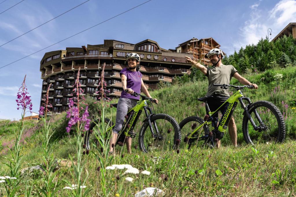 deux personnes avec des vélos dans un champ avec un bâtiment dans l'établissement Hôtel Les Menuires Les Bruyères, aux Menuires