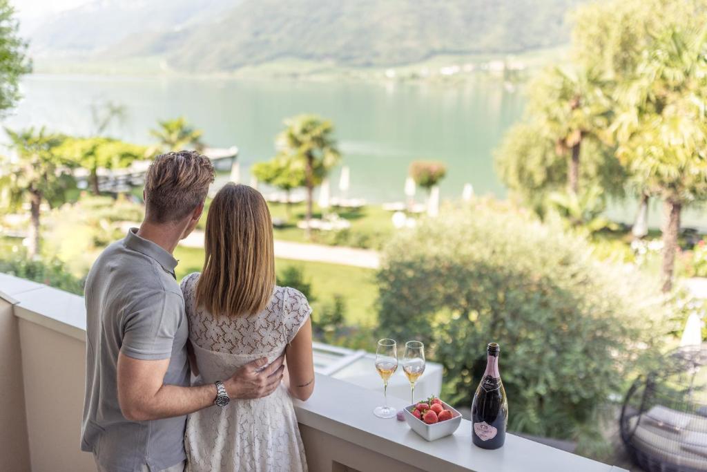 un homme et une femme debout sur un balcon avec des verres à vin dans l'établissement Parc Hotel Am See, à Caldaro