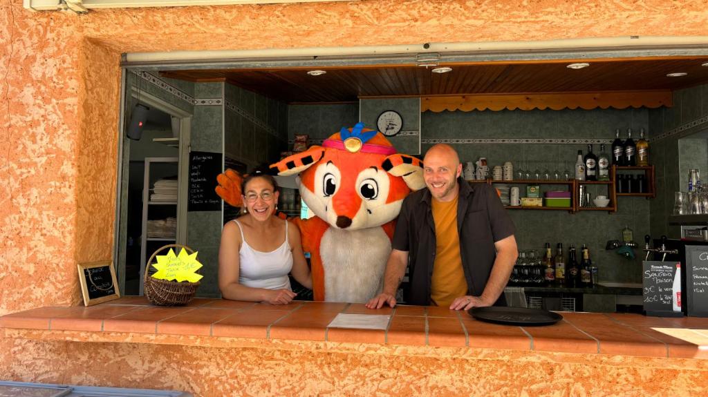 un homme et une femme debout derrière un bar avec une mascotte dans l'établissement Camping Tikayan L'Oxygène, à Valensole