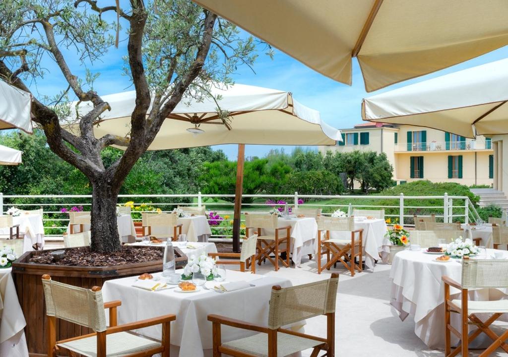 un restaurant avec des tables blanches et des parasols et un arbre dans l'établissement Tombolo Talasso Resort, à Marina di Castagneto Carducci