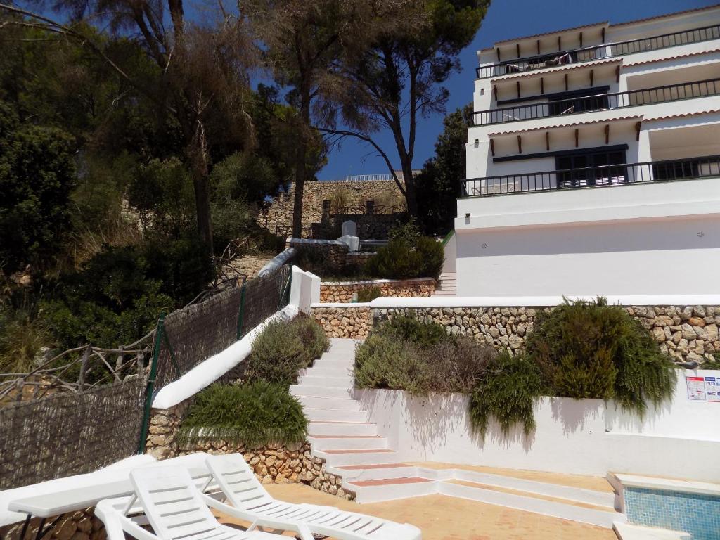 a building with white chairs and a swimming pool at Apartamentos Encanto Del Mar in Cala Galdana