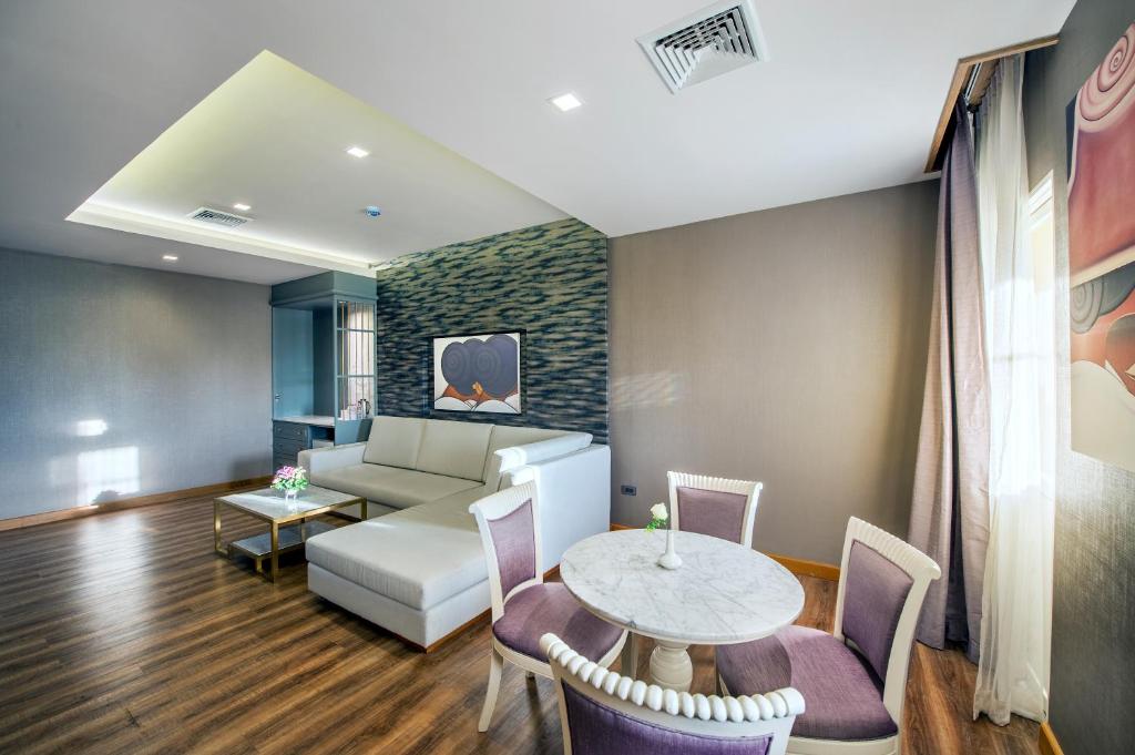 A bright suite sitting room with a sectional sofa, four-seat dining table and curtained windows.