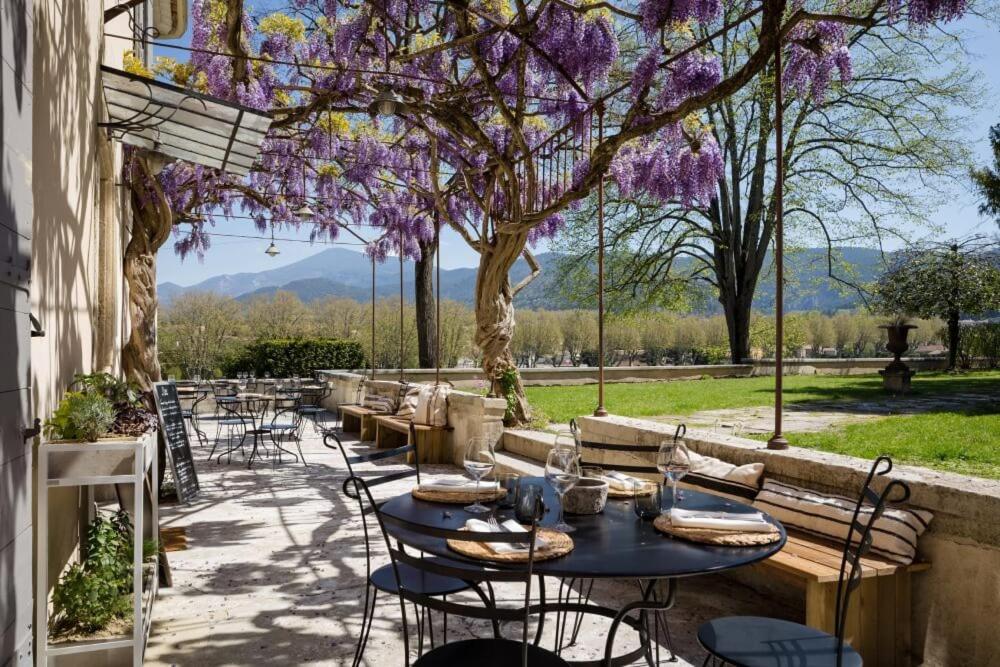 une terrasse avec des tables et des chaises sous un arbre et des fleurs violettes dans l'établissement Le Pont de L'orme, à Malaucène