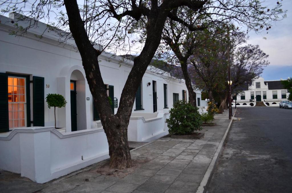 a white building with black windows and a tree on a street at De Kothuize 6 in Graaff-Reinet