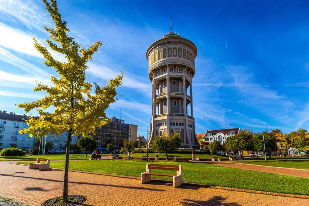 una torre en un parque con bancos y un árbol en Verona Apartman, en Szeged