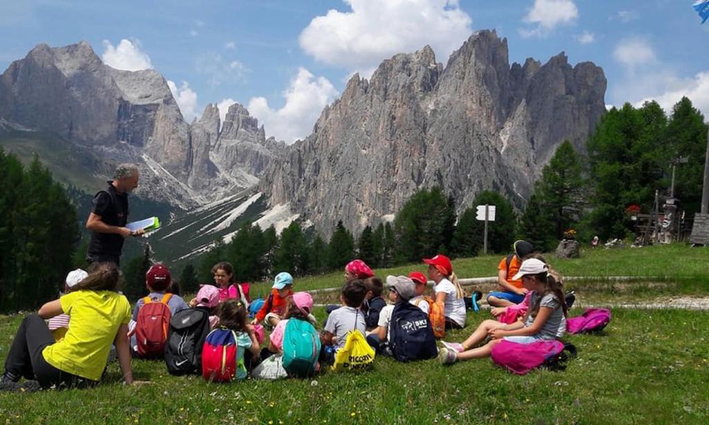 un gruppo di bambini seduti sul prato di fronte a una montagna di Family Hotel Gran Baita a Pozza di Fassa