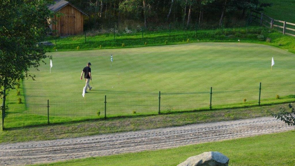 Golfer walking across fenced practice green, rustic shed and surrounding trees under soft sunlight.