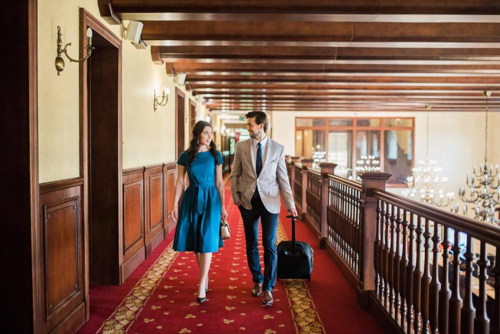 Couple walking down ornate hotel corridor, man pulling suitcase, cheerful atmosphere