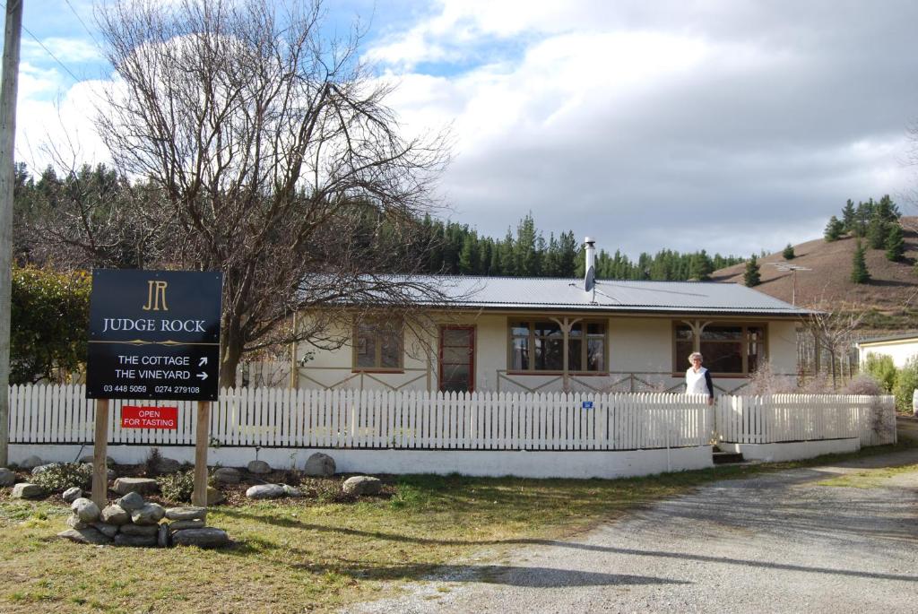 a woman standing in front of a house with a fence at Judge Rock Exclusive Vineyard Cottage Accommodation in Alexandra