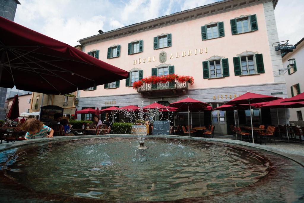 une fontaine en face d'un bâtiment avec des parasols rouges dans l'établissement Historic Hotel Albrici, à Poschiavo