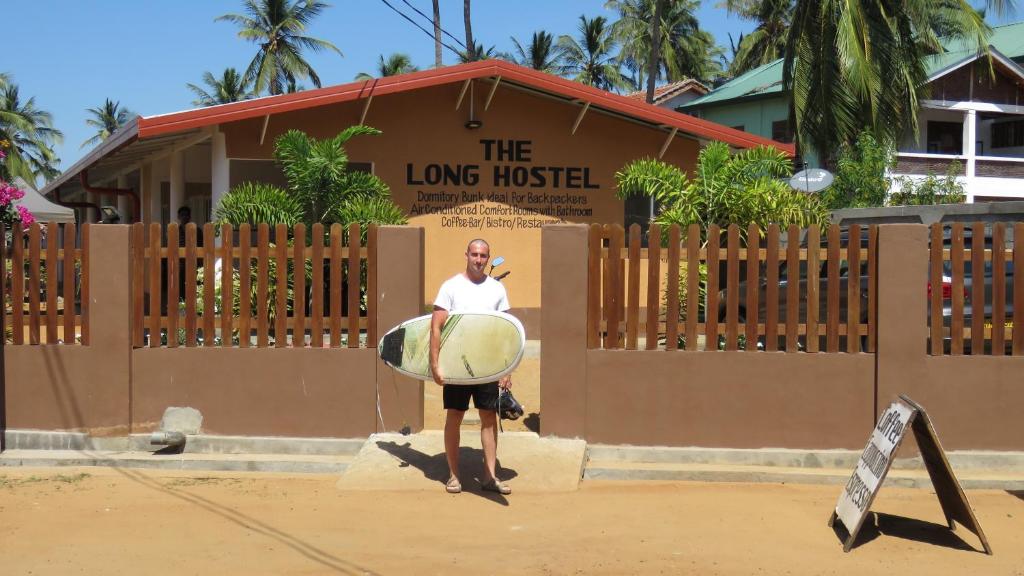 een man met een surfplank voor een gebouw bij The Long Hostel in Arugam Bay