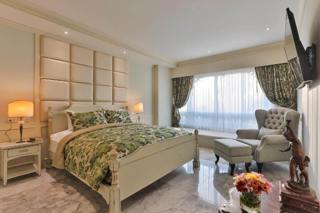 A traditional double bedroom with a padded headboard, large curtained window and lounge chair.