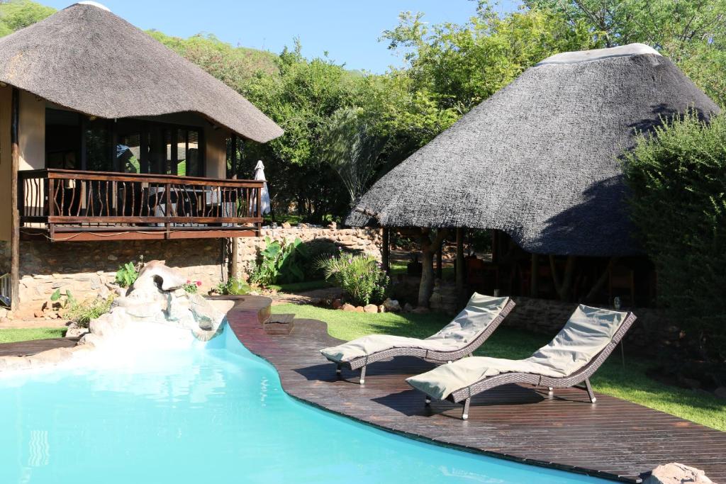 a swimming pool with two chairs and a thatch roof at Ijaba Lodge at Buschfeld Park in Outjo