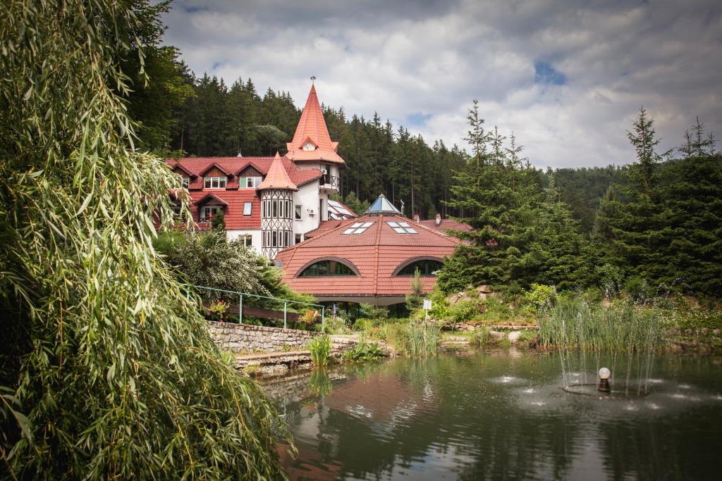 Resort building beside a reflective pond with fountain, framed by willow and forested hills.
