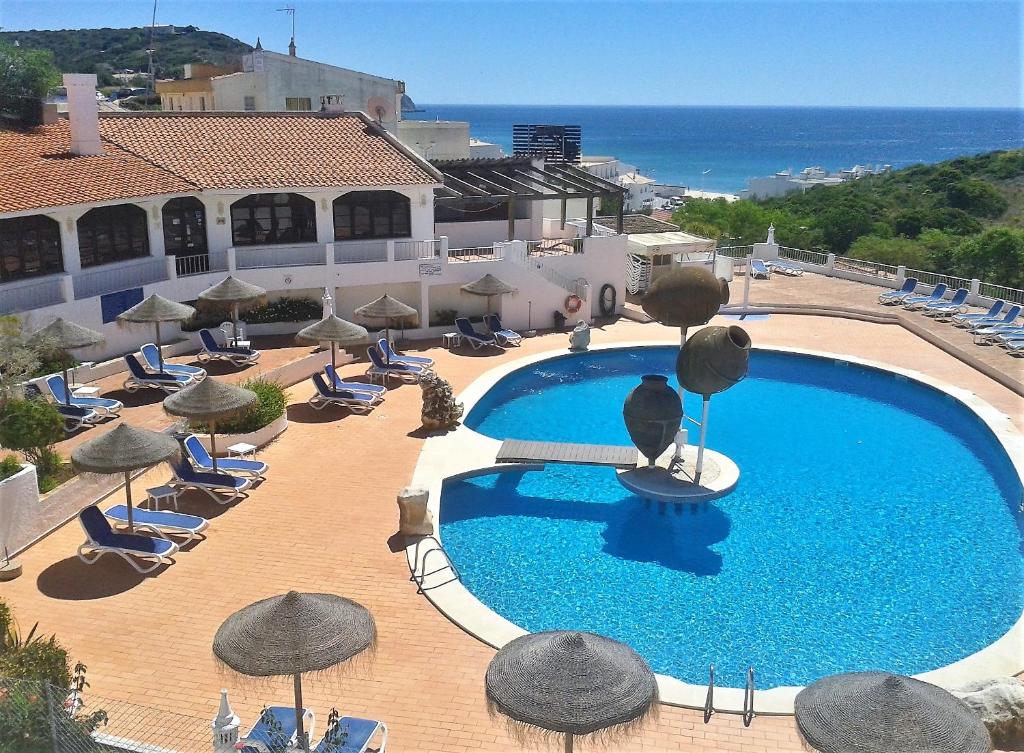 una piscina con sombrillas y sillas y el océano en Apartamentos Salema Praia Club, en Salema