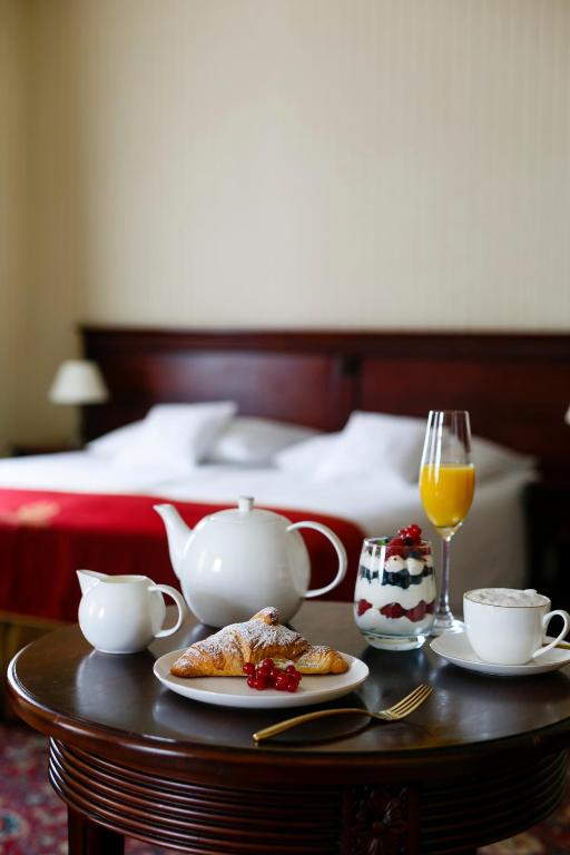 In-room breakfast on round table: croissant, teapot, orange juice, parfait, bed blurred in background.