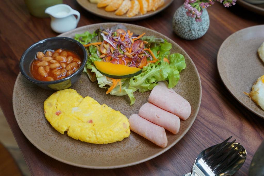 A plated cooked breakfast with omelette, sausages, salad, beans, and fruit nearby.