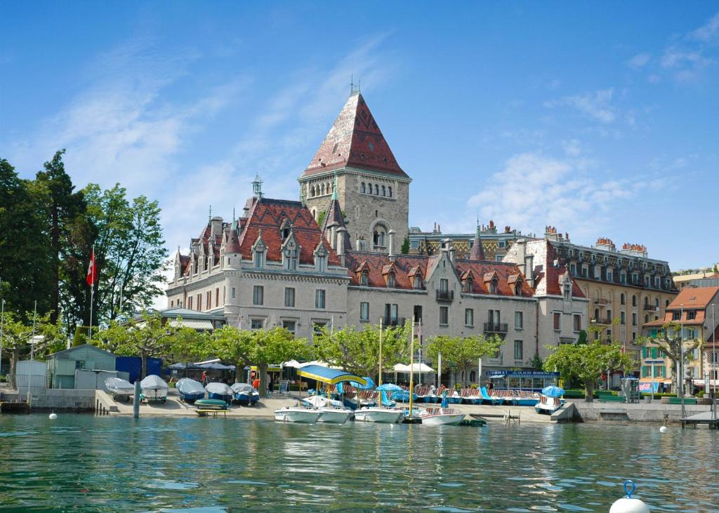 un grand bâtiment avec une tour d'horloge à côté d'une masse d'eau dans l'établissement Château d'Ouchy, à Lausanne