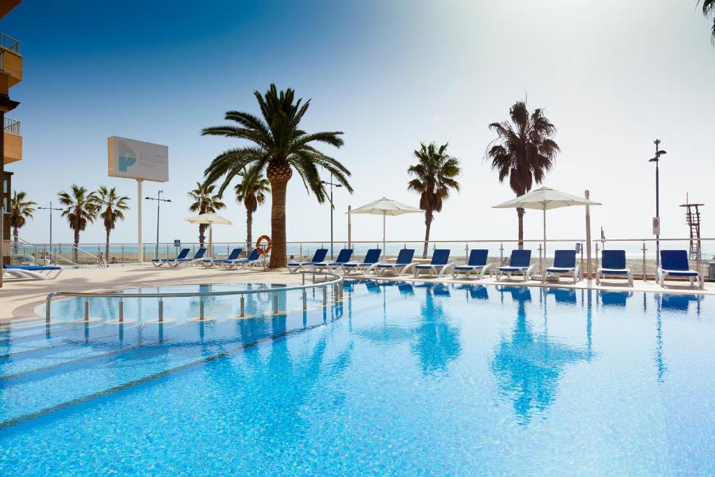 a swimming pool with chairs and palm trees and the ocean at Hotel Peñiscola Palace in Peniscola