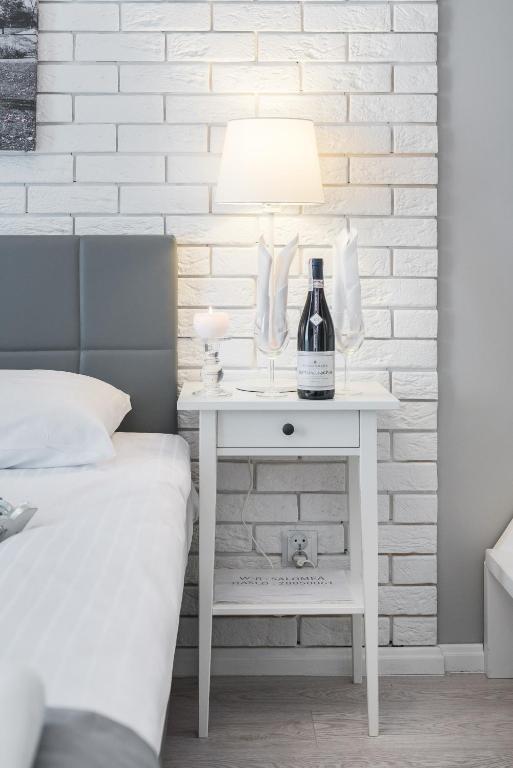 White bedside table with lit lamp, wine bottle and glasses against textured white brick wall.
