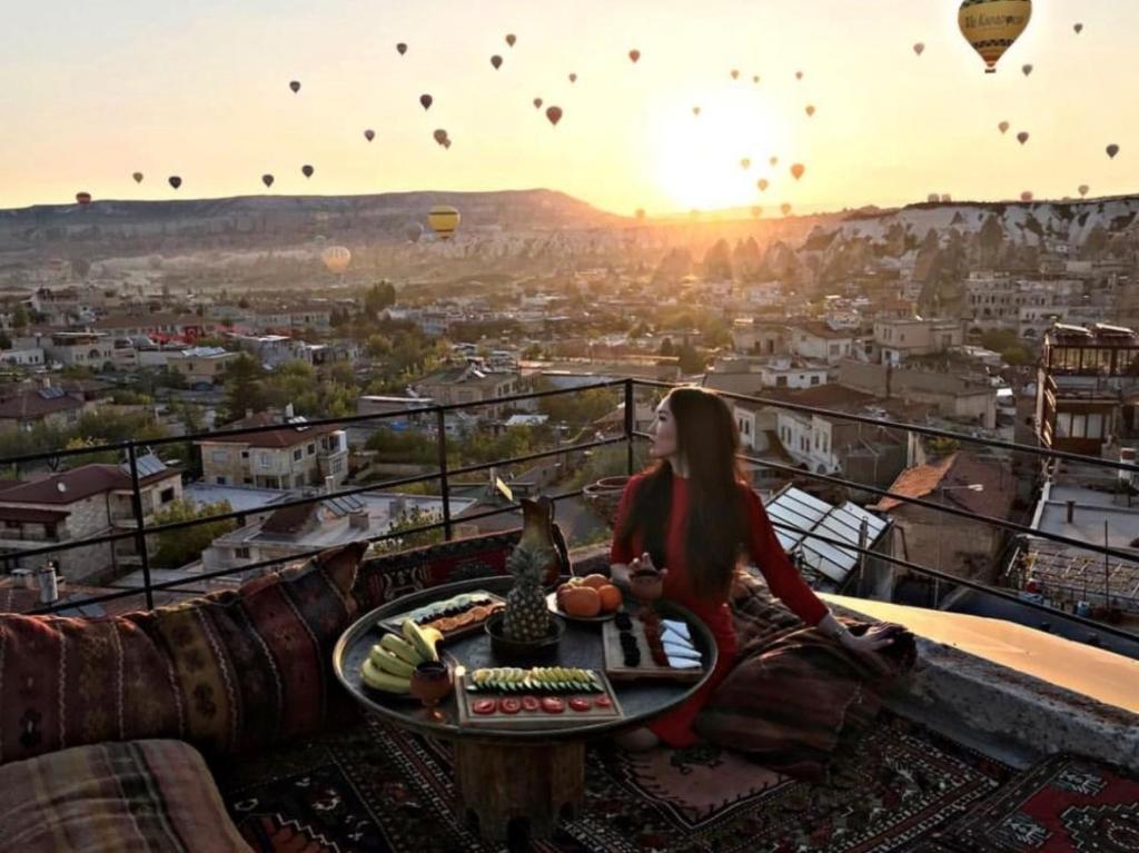 a woman sitting at a table on a balcony with hot air balloons at Travel Inn Cave Hotel in Göreme