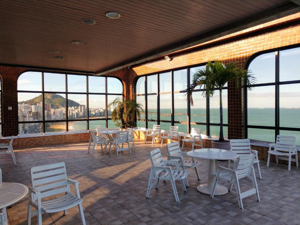 a restaurant with tables and white chairs and windows at Flat Pasárgada 712 - apt com vista para a Praia da Costa, academia moderna, piscinas panorâmicas, minimercado e lavanderia in Vila Velha