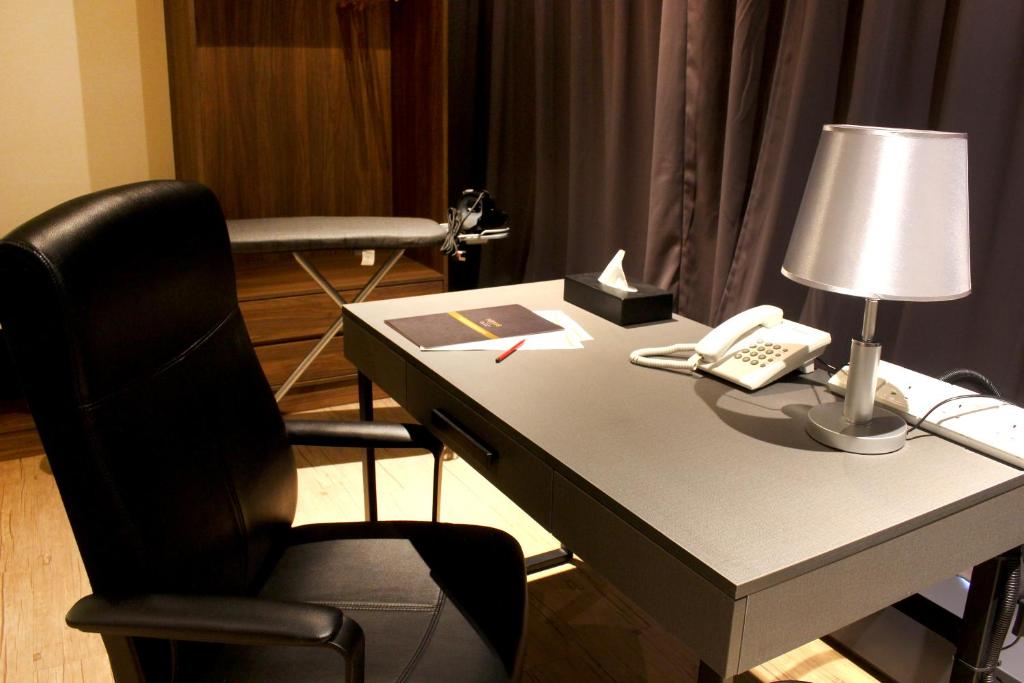 A furnished work desk with an office chair, lamp, telephone and ironing board behind it.