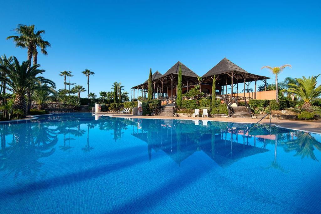 a large blue swimming pool with palm trees and huts at Puerto Antilla Grand Hotel in Islantilla