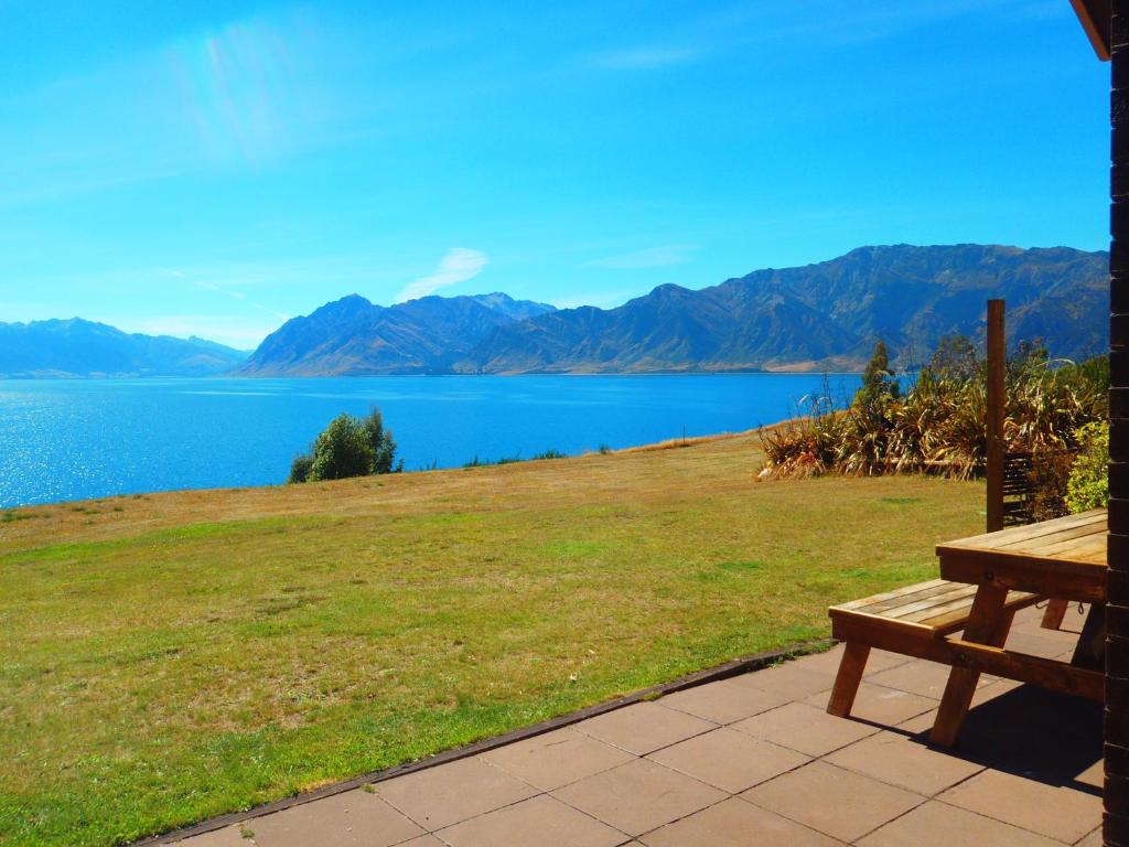 a wooden bench sitting on top of a field next to a lake at Lakefront Bellevue Lake Hawea Wanaka in Lake Hāwea