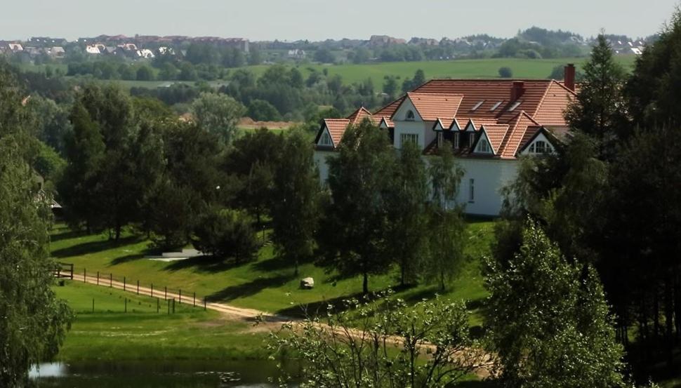 White country house with red roof nestled among trees on rolling green landscape, distant fields visible.