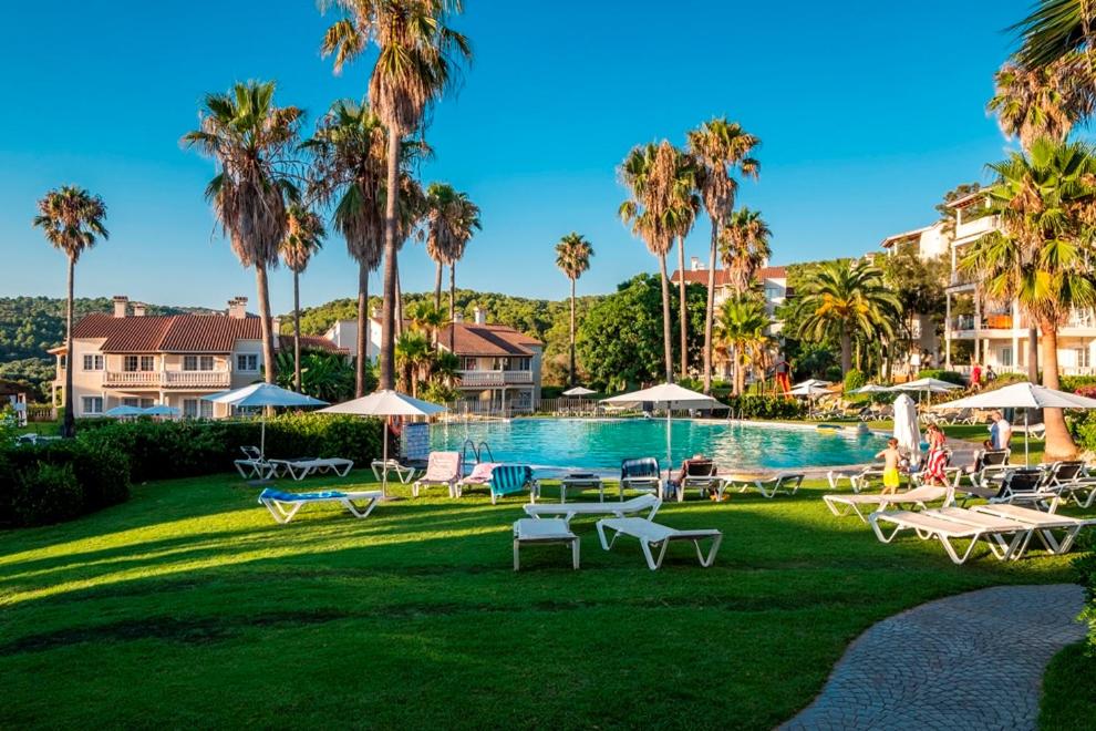 una piscina con sillas, sombrillas y palmeras en HG Jardín de Menorca, en Son Bou