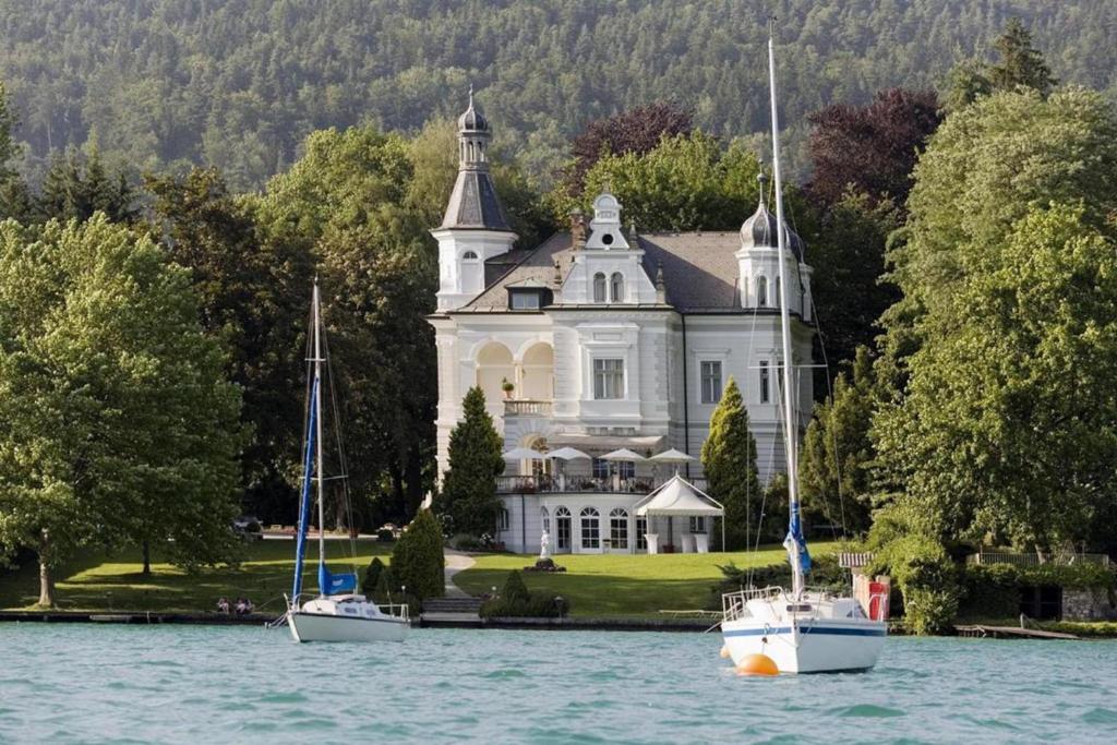 due barche sull'acqua di fronte a una grande casa di Dermuth Hotels – Parkvilla Wörth a Pörtschach am Wörthersee