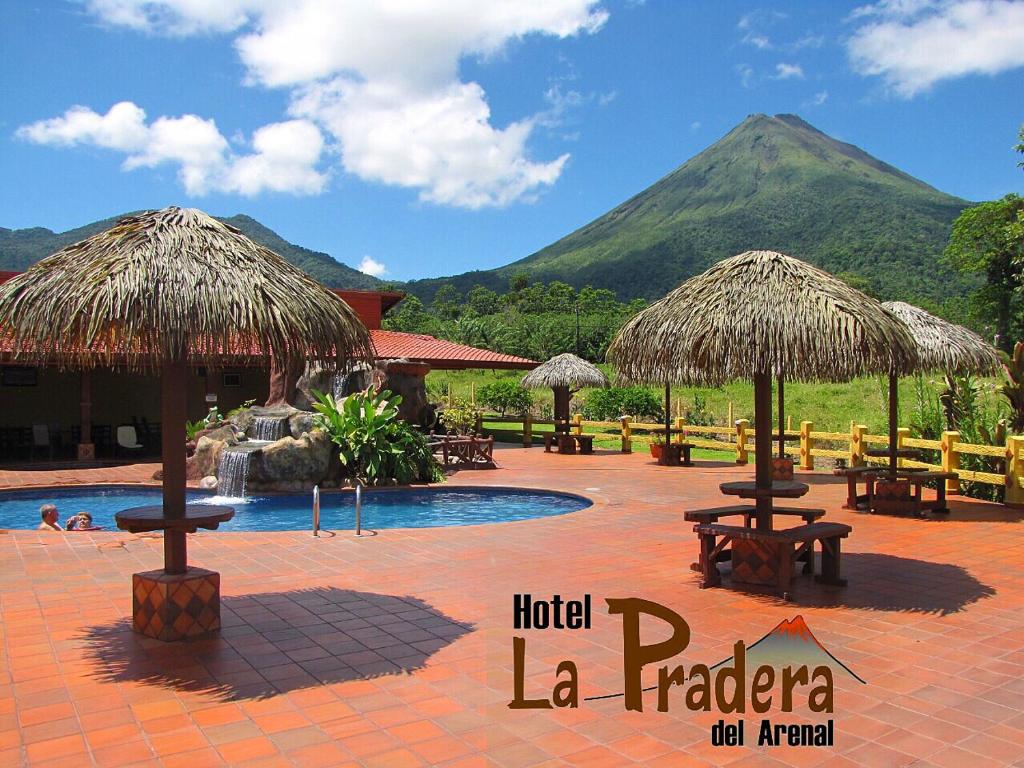 a resort with a pool with tables and straw umbrellas at Hotel La Pradera del Arenal in Fortuna