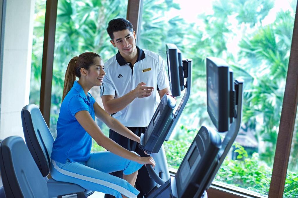 A bright fitness room has cardio machines beside large windows overlooking greenery.