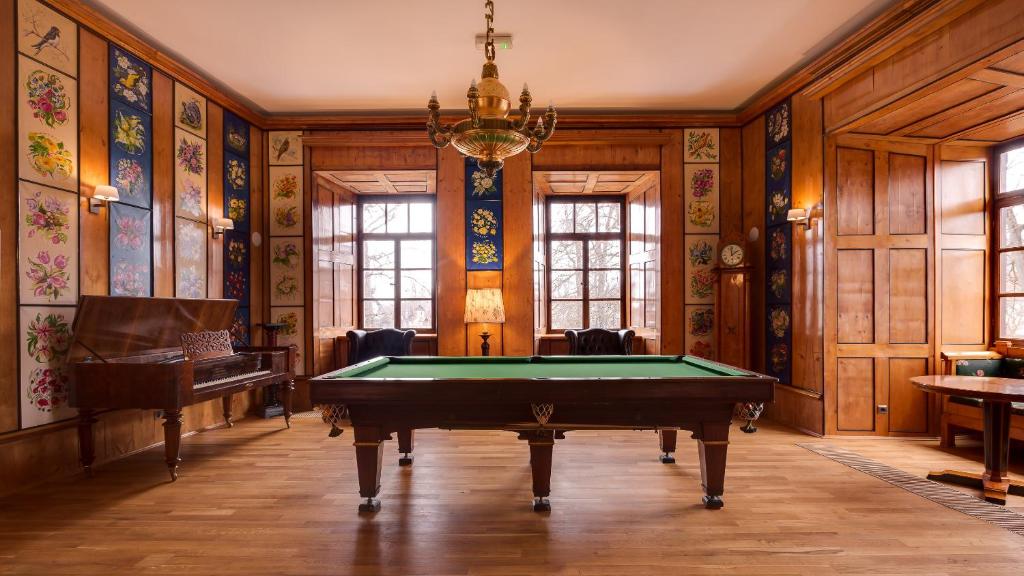 Elegant billiards room with green pool table, decorative floral wall panels, piano, tall windows.