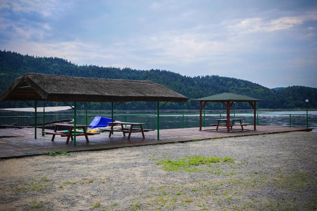 Quiet lakeside pier with covered picnic tables, small boats and tree-covered hills in background.