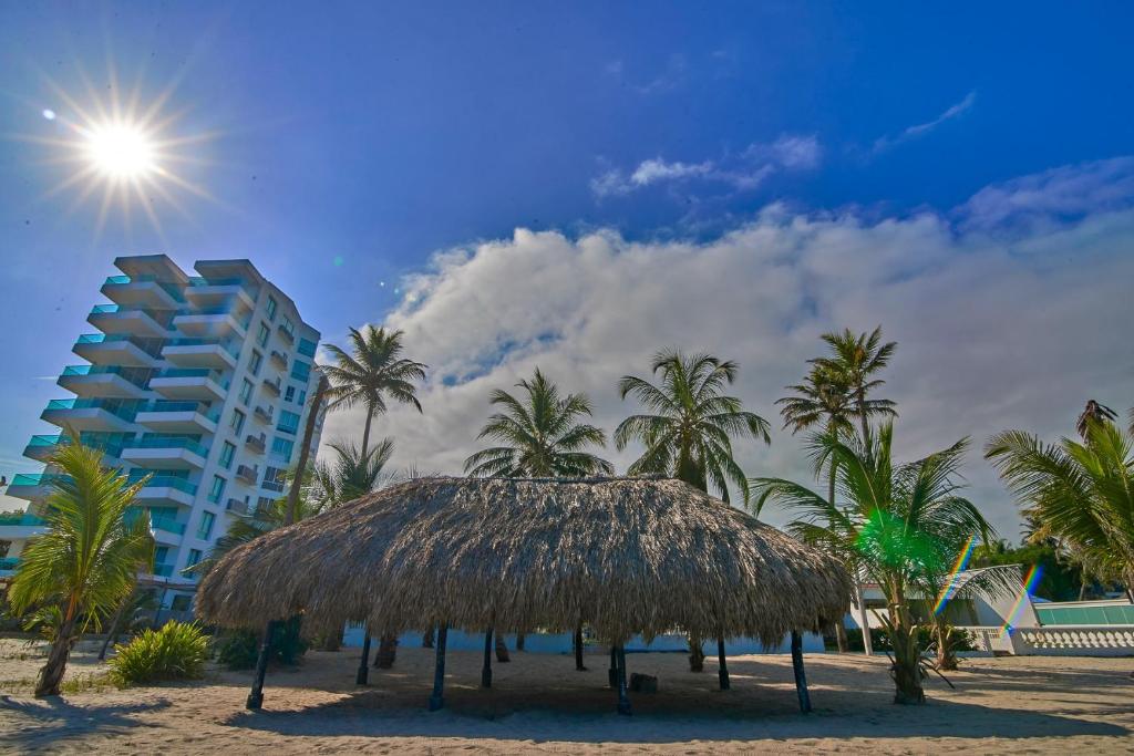 a straw hut on a beach with palm trees and a building at Cabaña Coveñitas 2 in Coveñas