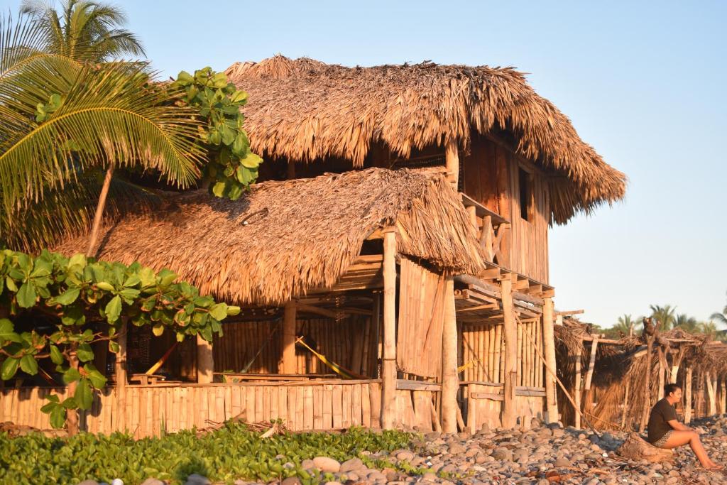 un hombre sentado frente a una cabaña de paja en Pelicano Surf Camp en La Libertad