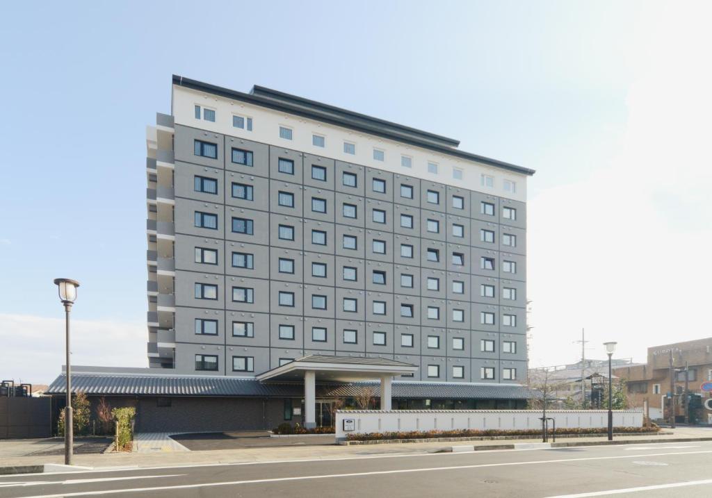 um edifício cinza alto com uma paragem de autocarro em frente a uma rua em Hotel Route Inn Kasai Hojonoshuku em Kasai