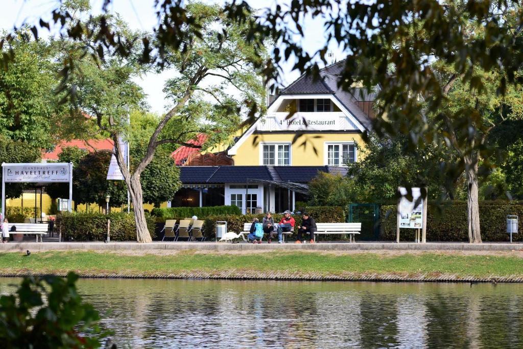 uma casa amarela com pessoas sentadas em bancos ao lado de um lago em Apart Hotel Haveltreff em Caputh