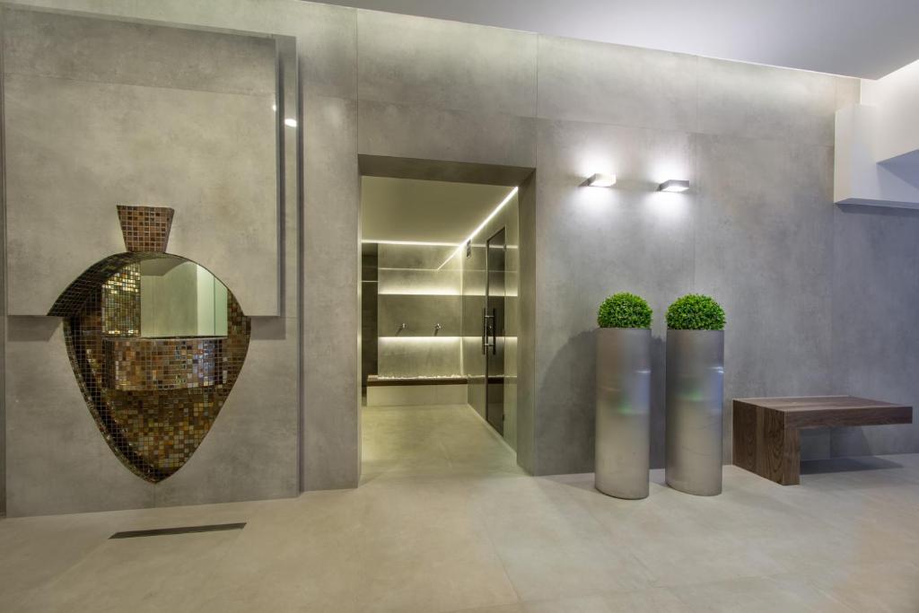 Minimal concrete spa corridor with mosaic vase niche, two tall planters, wooden bench, soft lighting.