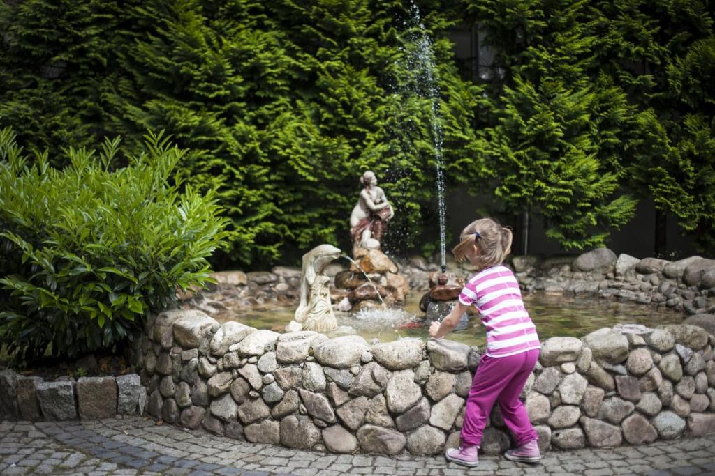 Child leaning over stone fountain with water jets and small statues, playful garden moment
