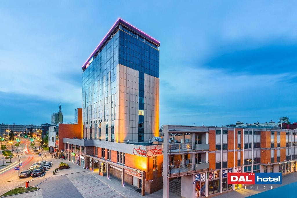 Modern glass-front hotel tower lit at dusk, street-level shops, parked cars and hotel signage visible.