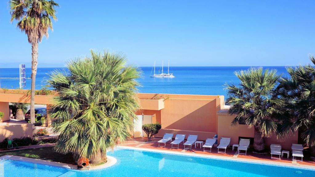 a resort with a swimming pool with palm trees and the ocean at Hotel Punta Nord Est in Castellammare del Golfo