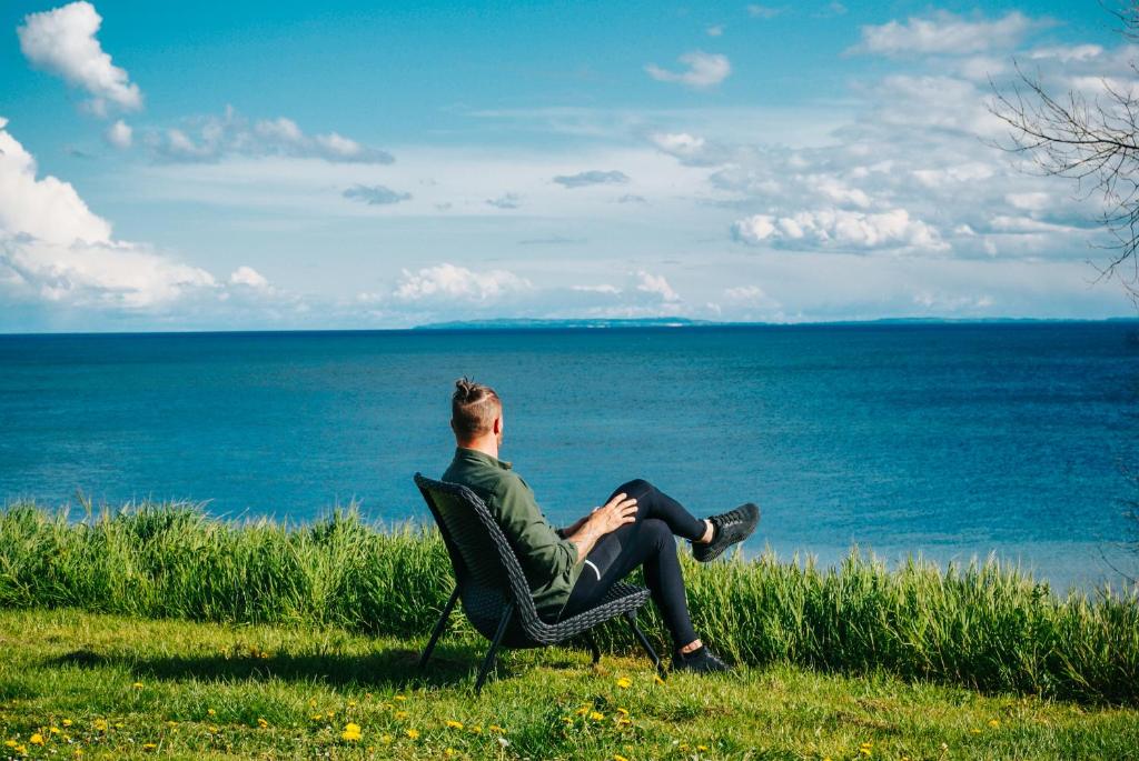 a man sitting in a chair looking out at the ocean at Recharging luxury stay for maksimum 4 people with magnificent sea view NO BREAKFAST - One bathroom - Hottub open from 15April-1November in Fakse Ladeplads