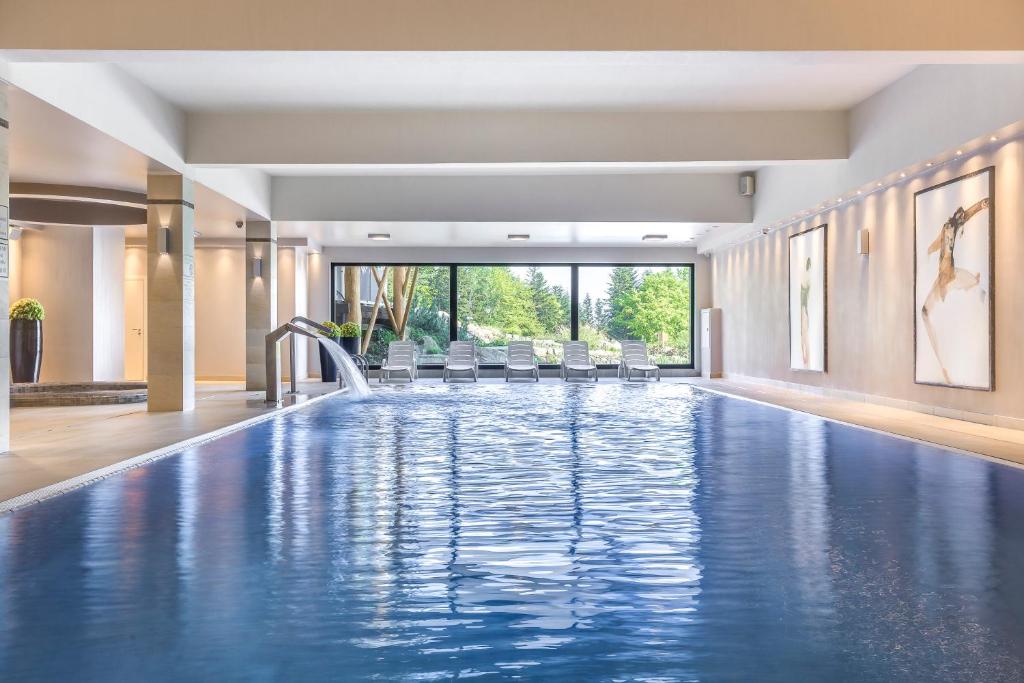 Bright indoor swimming pool with water cascade, row of lounge chairs facing large garden windows.